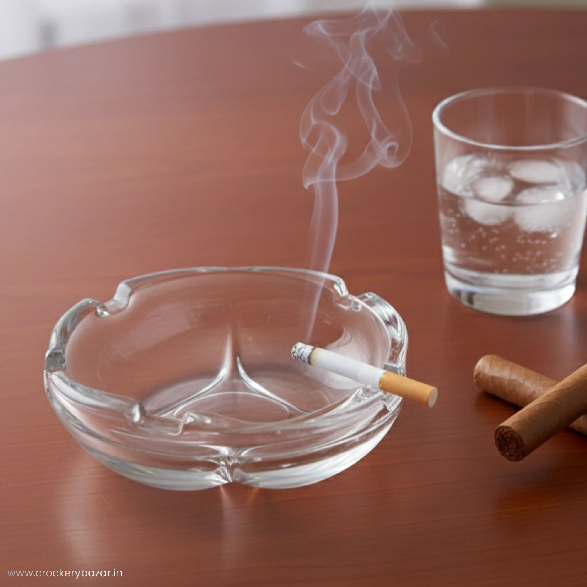 A small clear glass ashtray on a modern wooden office desk next to a glass of water and cigars.