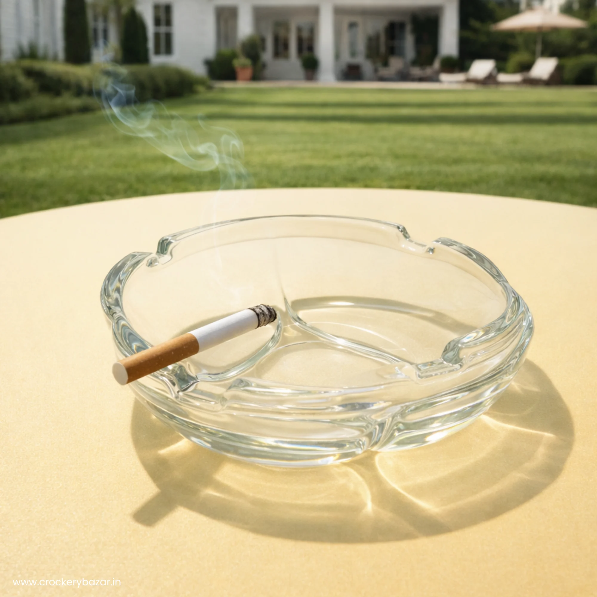 A clear glass ashtray on a yellow outdoor table in a garden setting with a smoking cigarette.