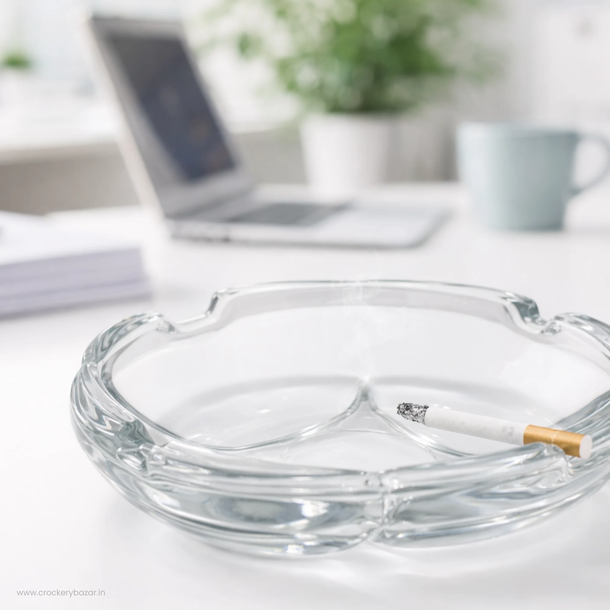 A clear glass ashtray on a white office desk next to a laptop and a blue coffee mug.