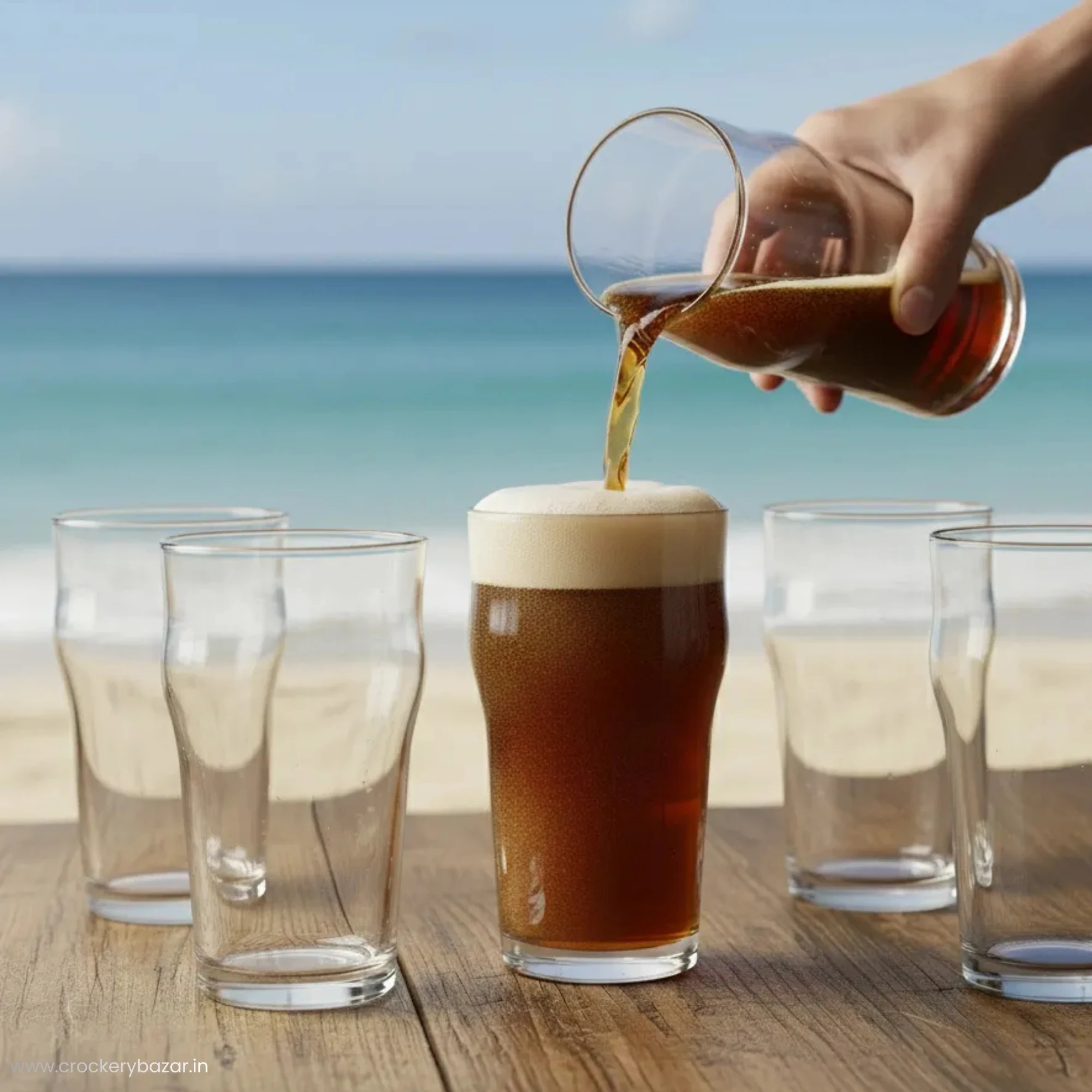A hand pouring a dark stout beer into a Nonic glass on a wooden table with a beach background.