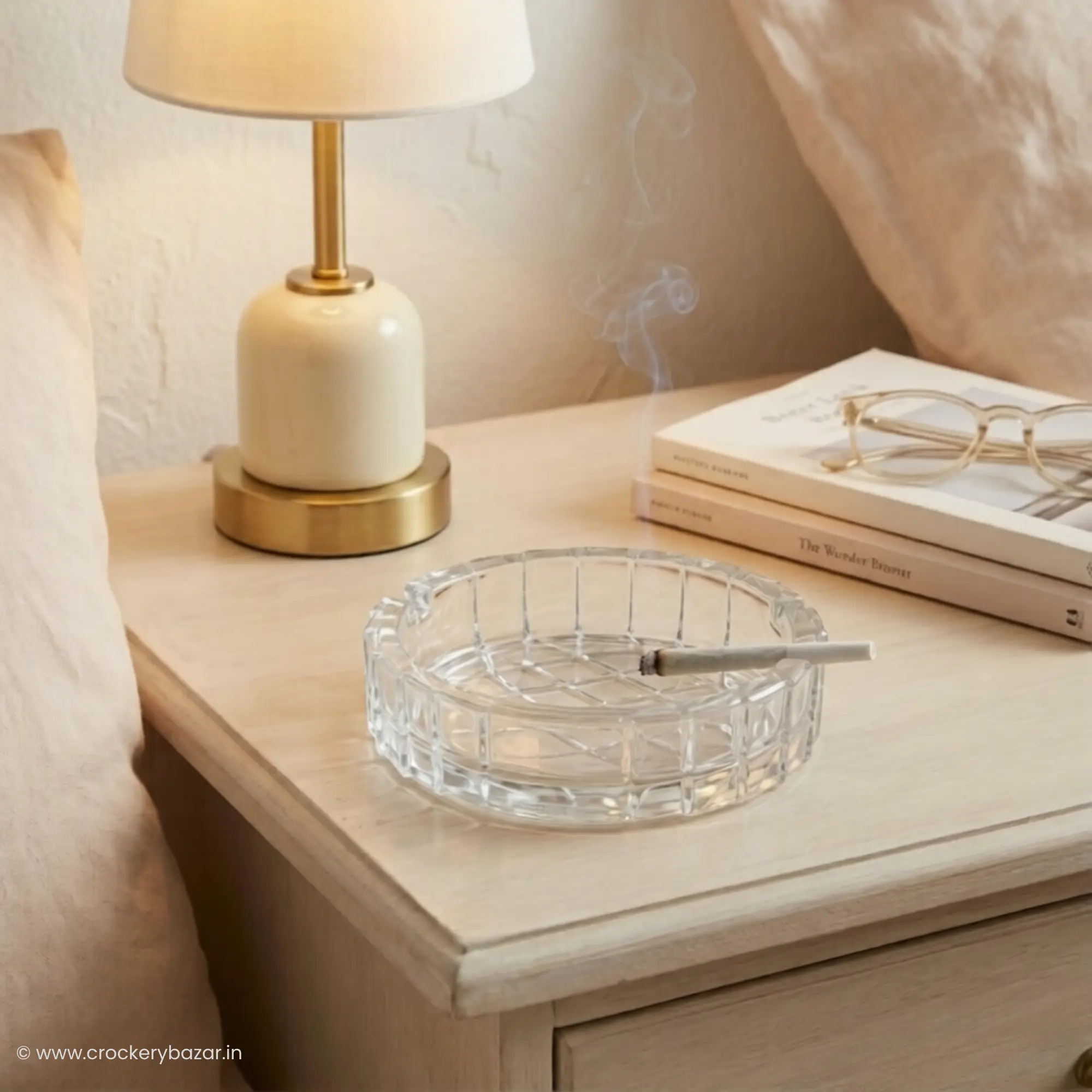 A Bormioli Rocco Duemila large glass ashtray placed on a light-colored wooden bedside table next to a stylish lamp, books, and eyeglasses.