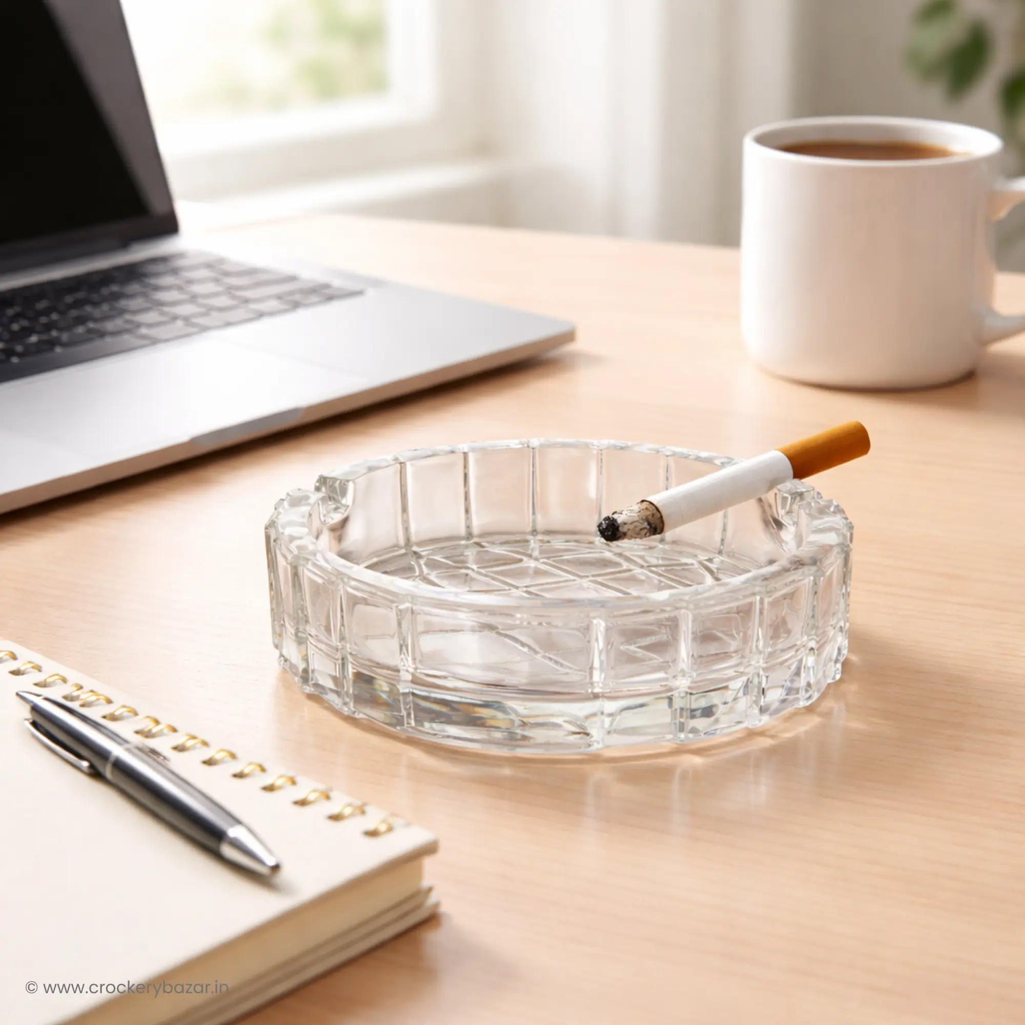 A Bormioli Rocco Duemila big size ashtray sitting on a light wood desk next to a laptop, a notebook, and a white coffee mug in a professional office setting.